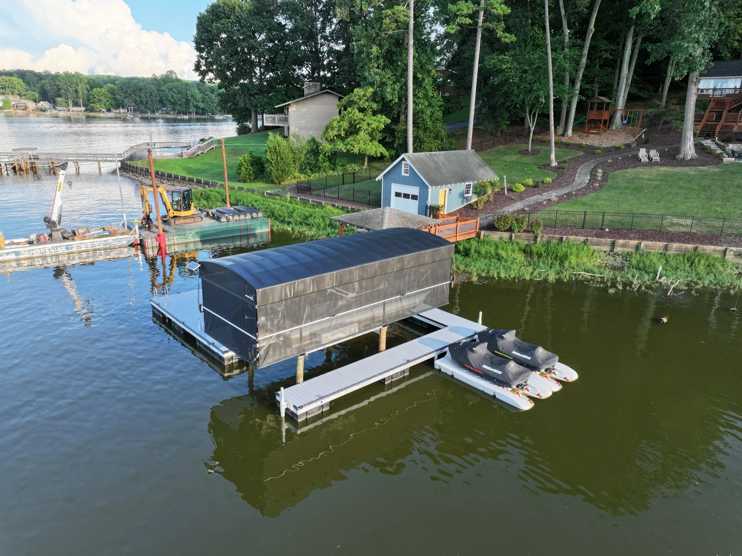 A marina by a lake showing a dock with boats, a small blue shed, a paved pathway, trees, and a house on a hill in the background.