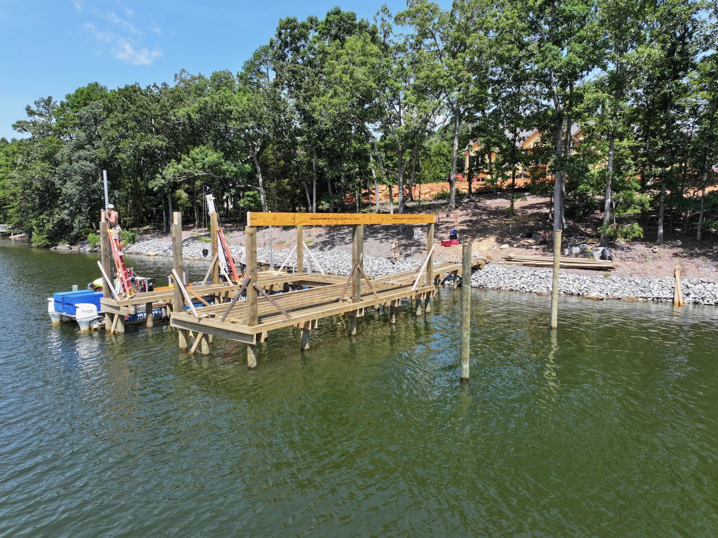 Construction workers building a wooden dock or pier on a lake, surrounded by green trees and a house in the background.