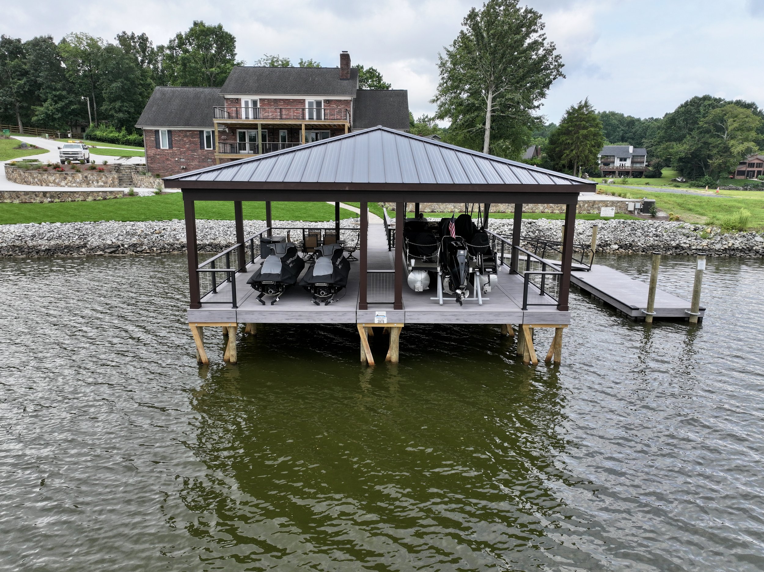 A lakeside scene with a wooden dock and covered boat storage area, boats and jet skis parked underneath, a stone retaining wall, a brick house with a deck, green lawn, trees, and neighboring houses in the background.