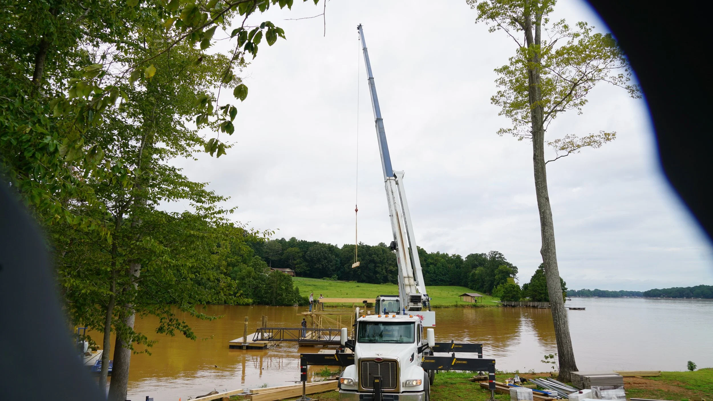 A crane with an extended boom is building a dock or pier by a body of water surrounded by trees, with a cloudy sky overhead.