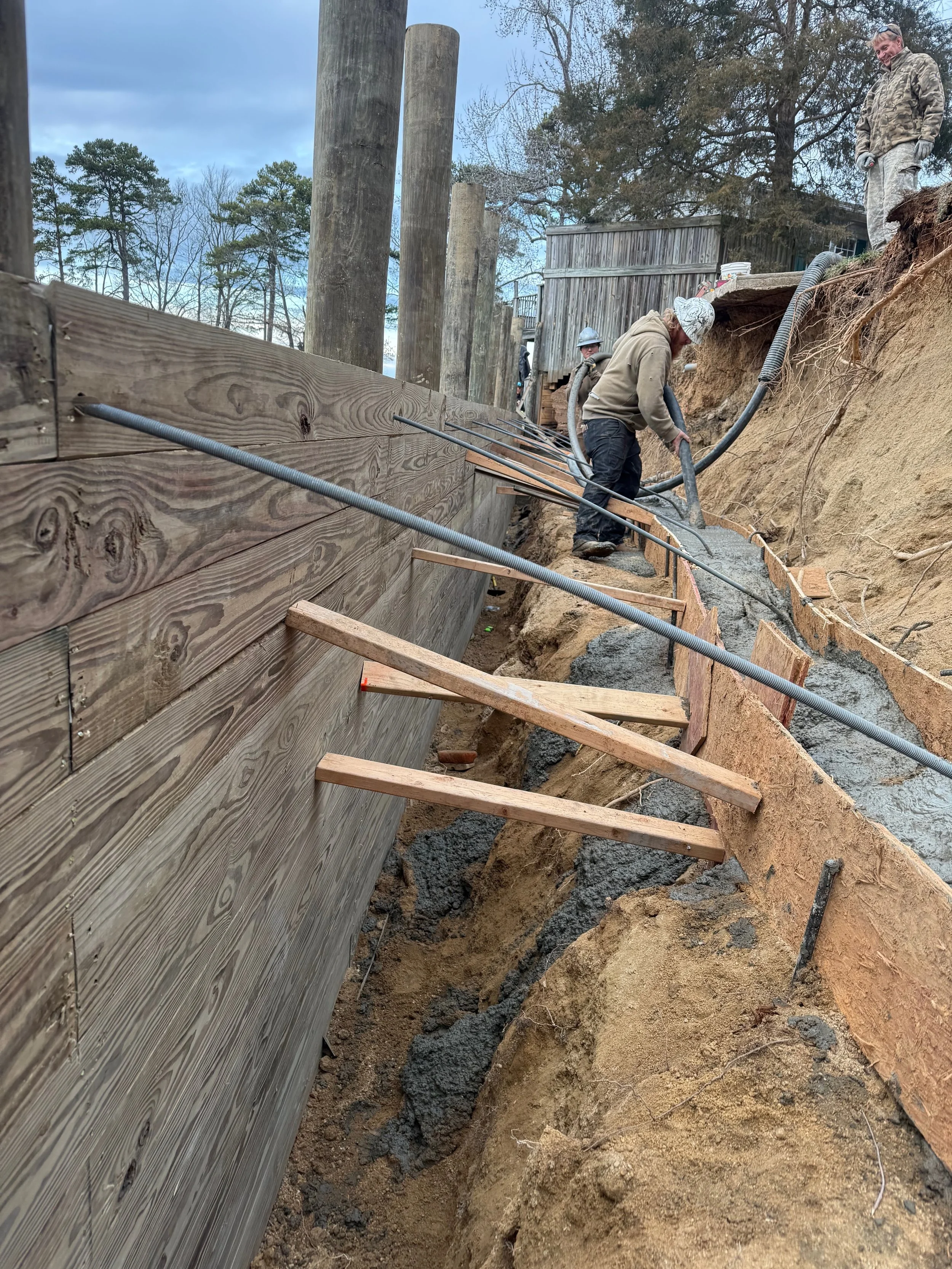 Construction workers laying concrete in a trench beside a wooden seawall, with trees in the background.