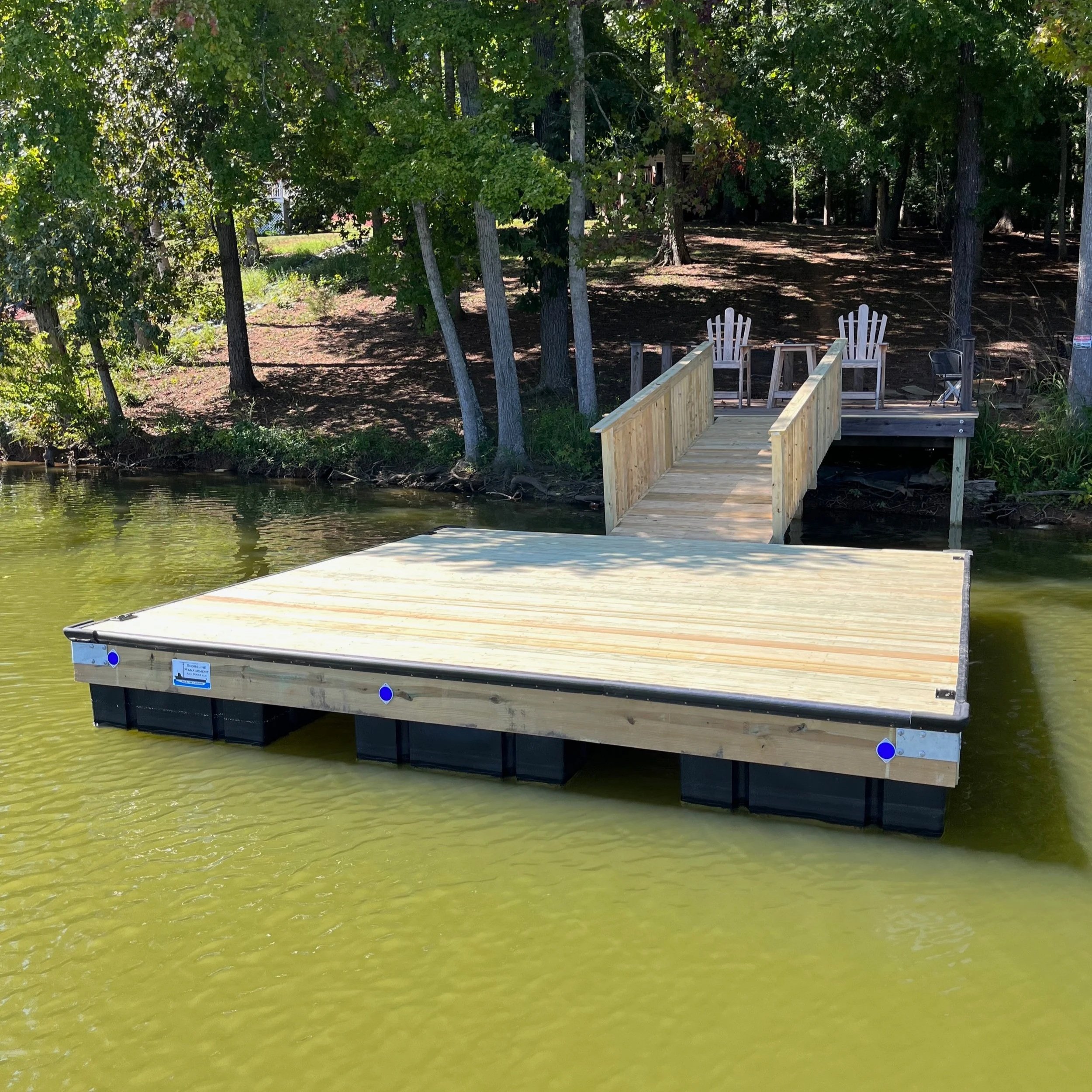 A wooden dock extends into a calm greenish lake, surrounded by lush green trees.