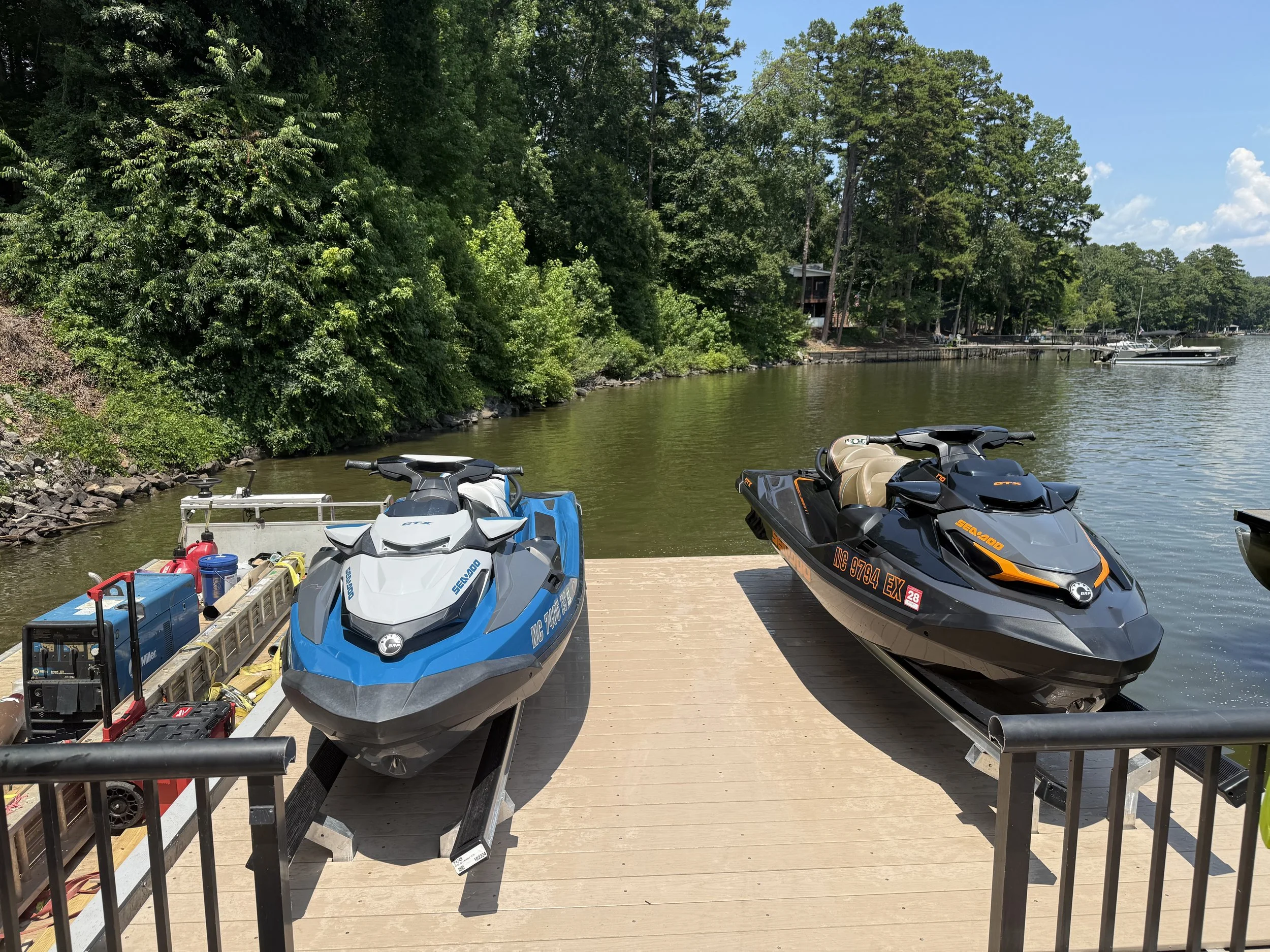 A dock with four personal watercraft (Jet Skis) parked on it, next to a lake with trees along the shoreline and boats in the water, under a blue sky with some clouds.