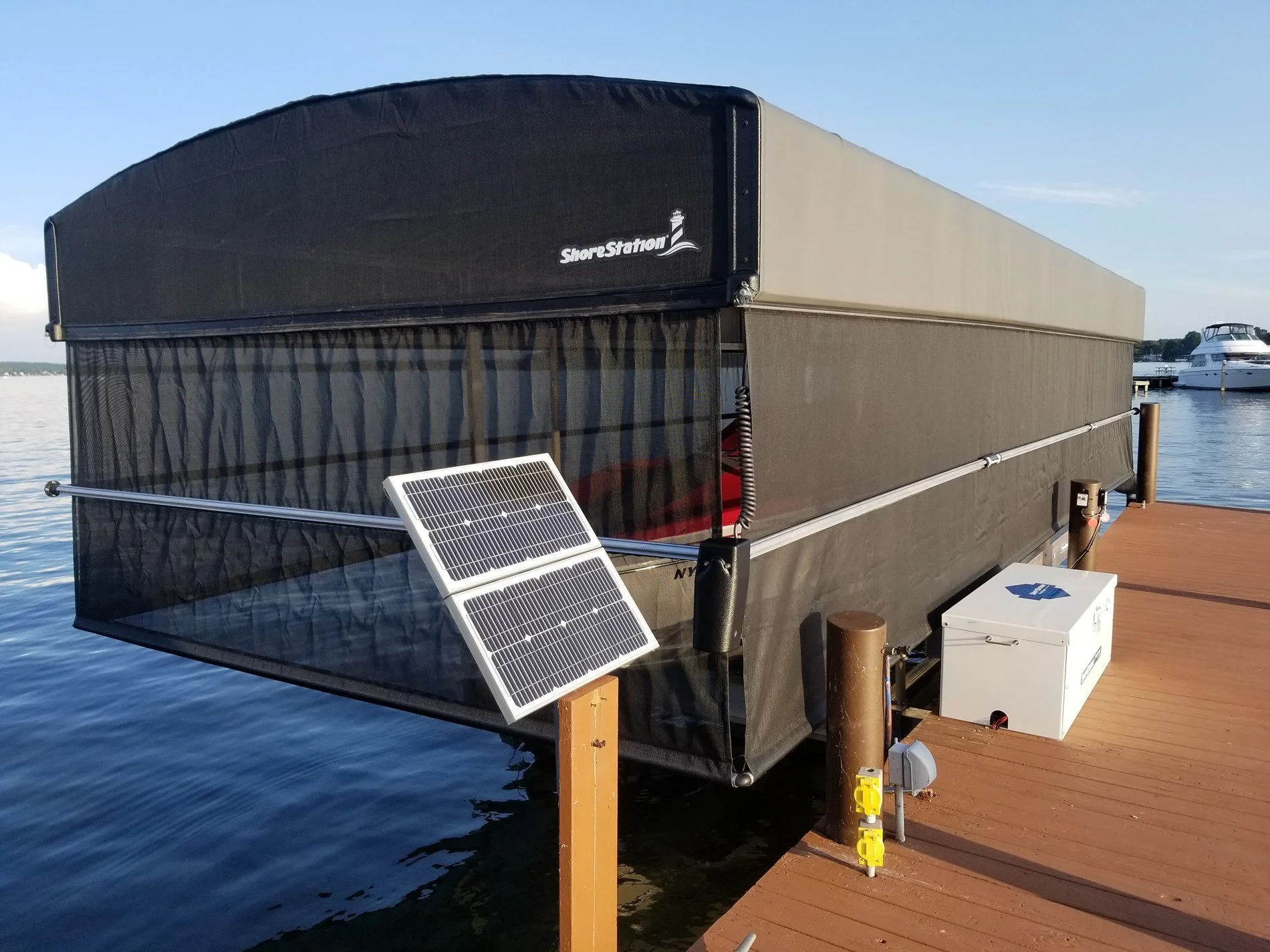 A floating dock with solar panels, a small white utility box, and a black ShoreStation boat lift on a body of water, with a boat in the background.