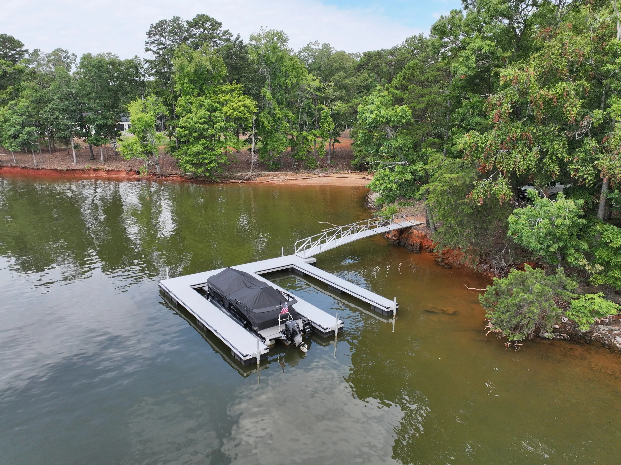 A boat docked at a marina in a lake, with lush green trees on the shoreline and a small sandy beach area, under a cloudy sky.