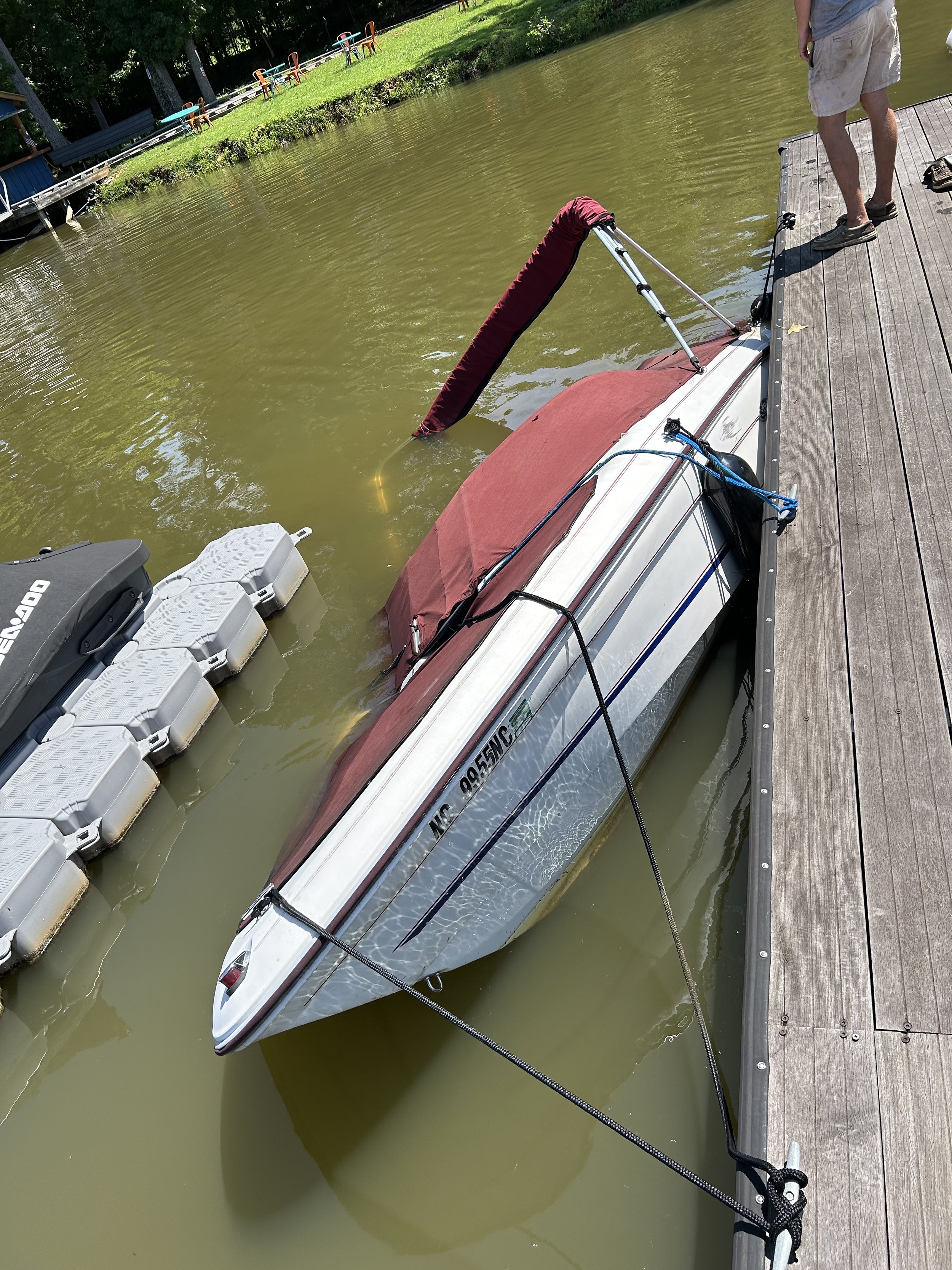 A small boat is partially submerged in murky green water next to a wooden dock. A person is standing on the dock, and a tarp covers part of the boat.