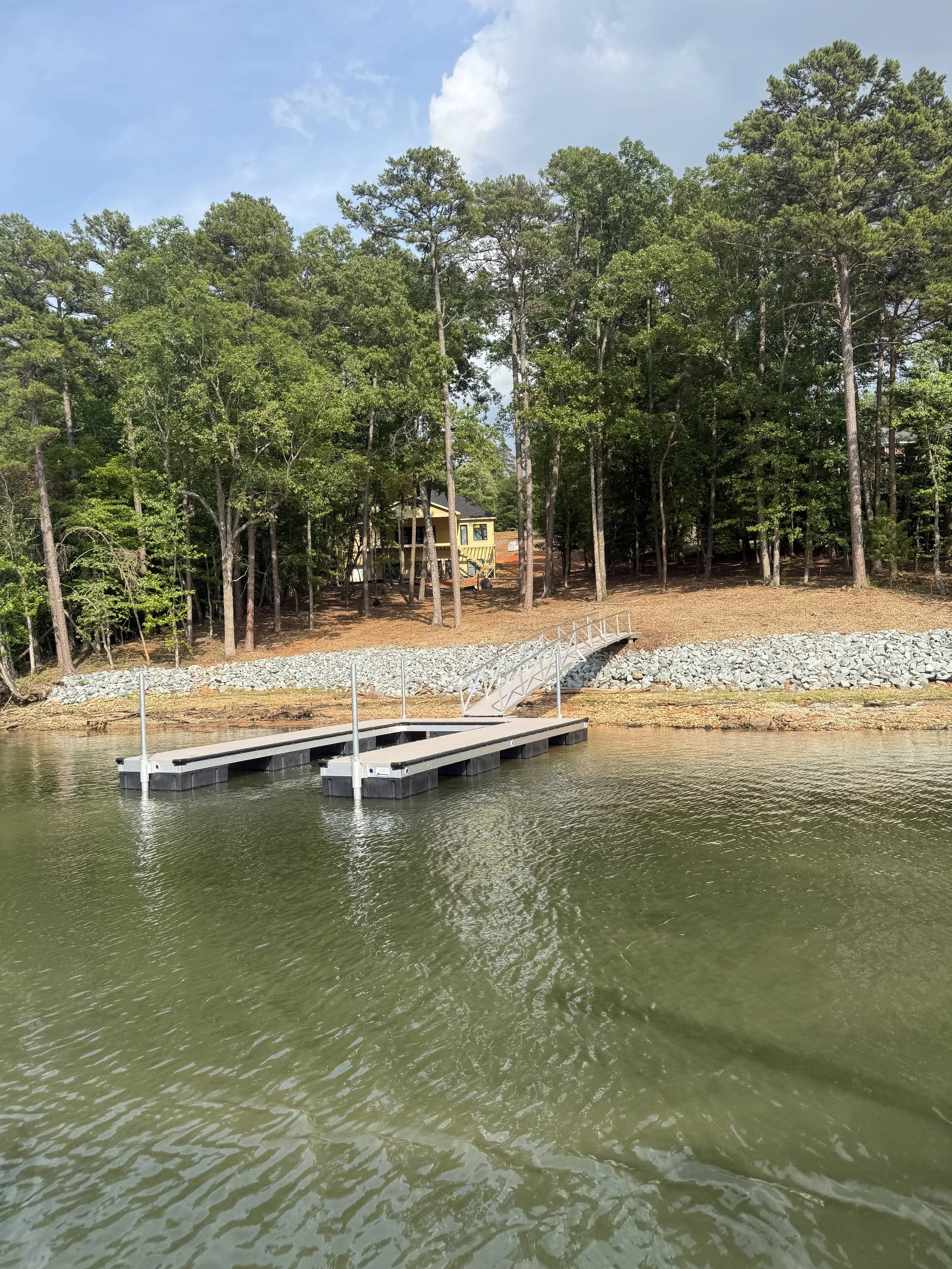 A small dock extends into a body of water with a forested hillside in the background, featuring tall trees and a partly cloudy sky.