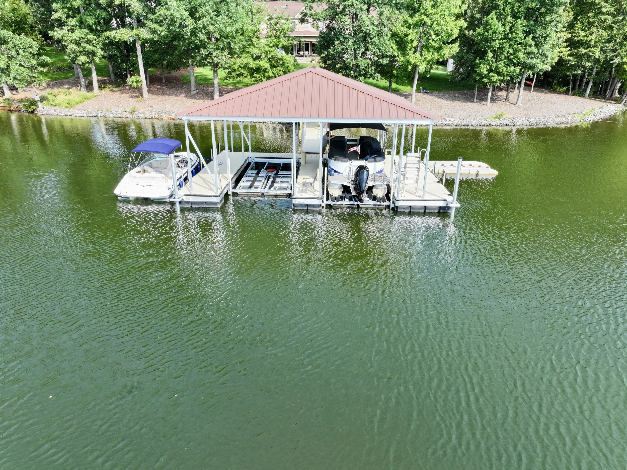 A boat dock with a red roof on a lake, surrounded by green trees and a house in the background.