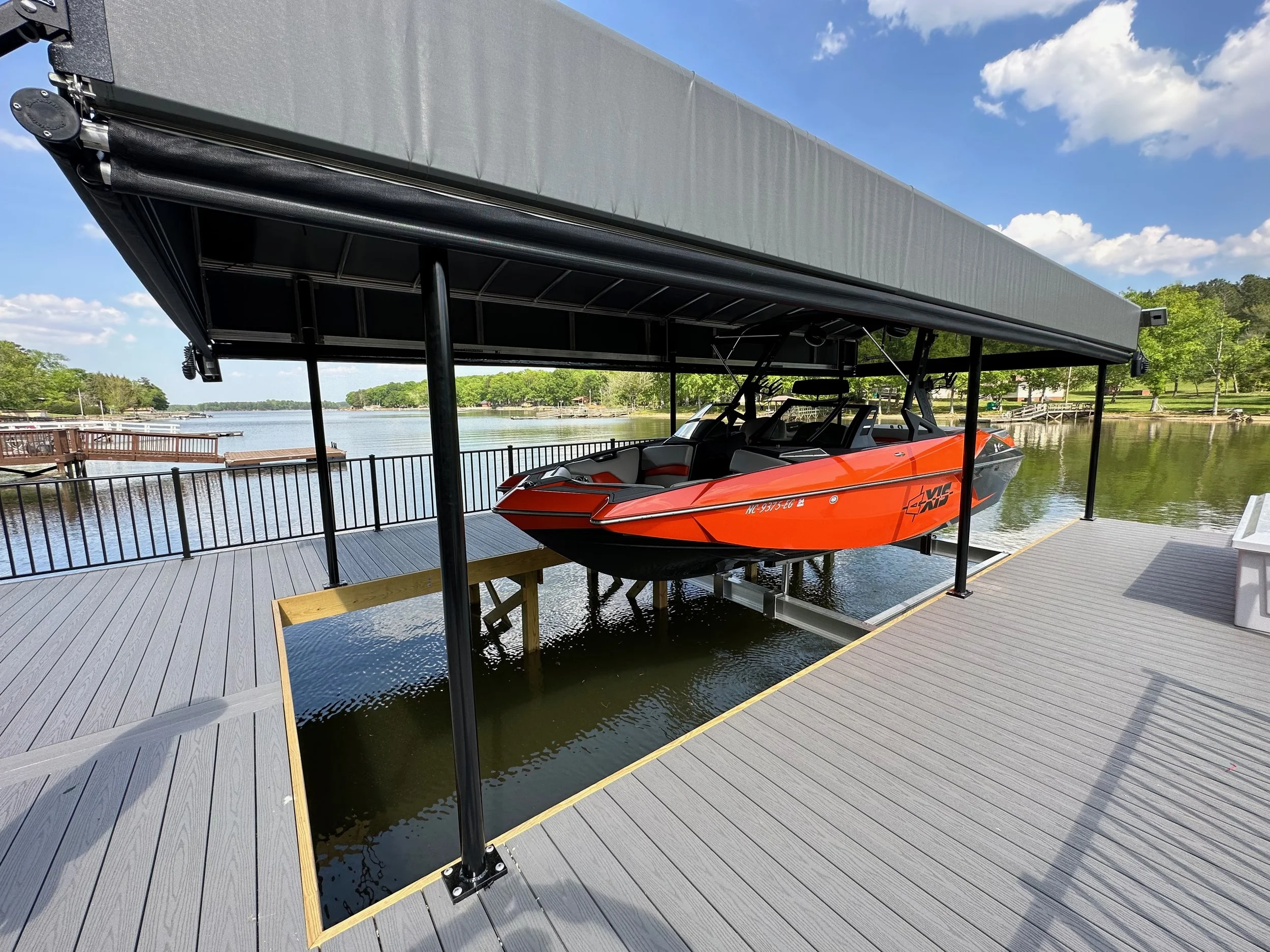 A red and black speedboat parked on a floating dock by a lake, under a boat cover with a cloudy blue sky in the background.