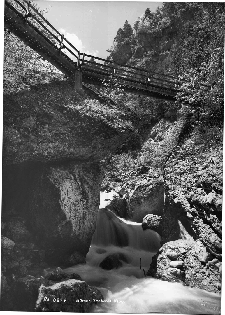 Schwarz-weiß-Foto von einem Wasserfall in einer felsigen Schlucht, über der eine Brücke mit Geländer führt, umgeben von Bäumen und Felsen.