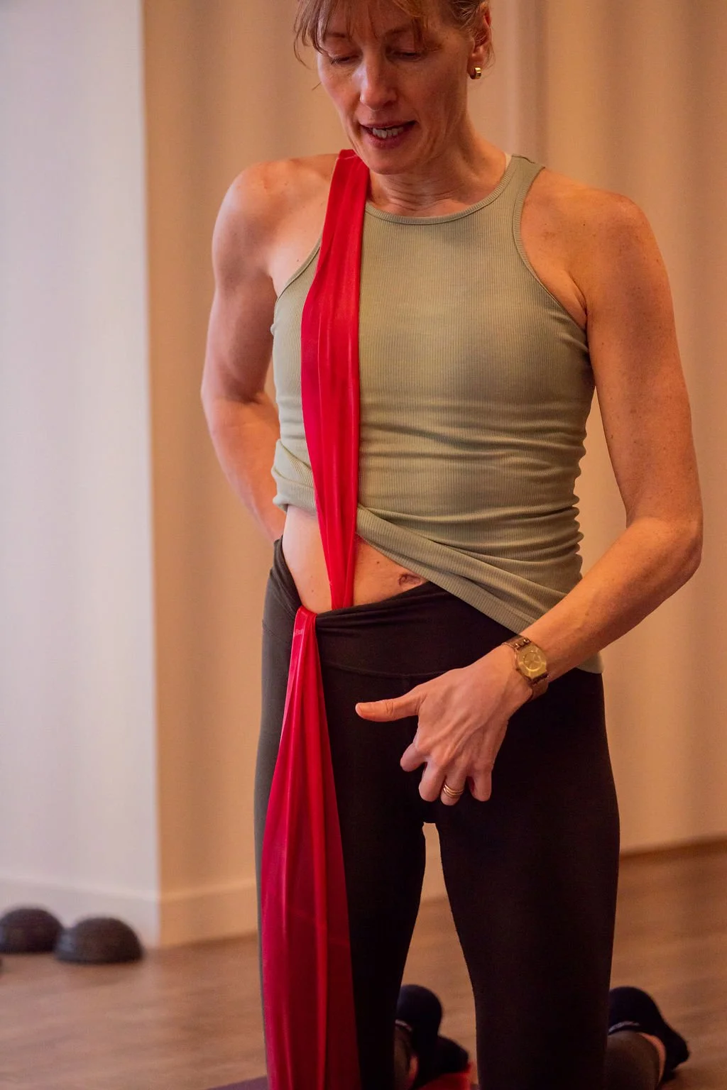 A woman with short hair wearing a beige tank top, black pants, and a red scarf around her neck, holding her lower abdomen and pointing with her finger.