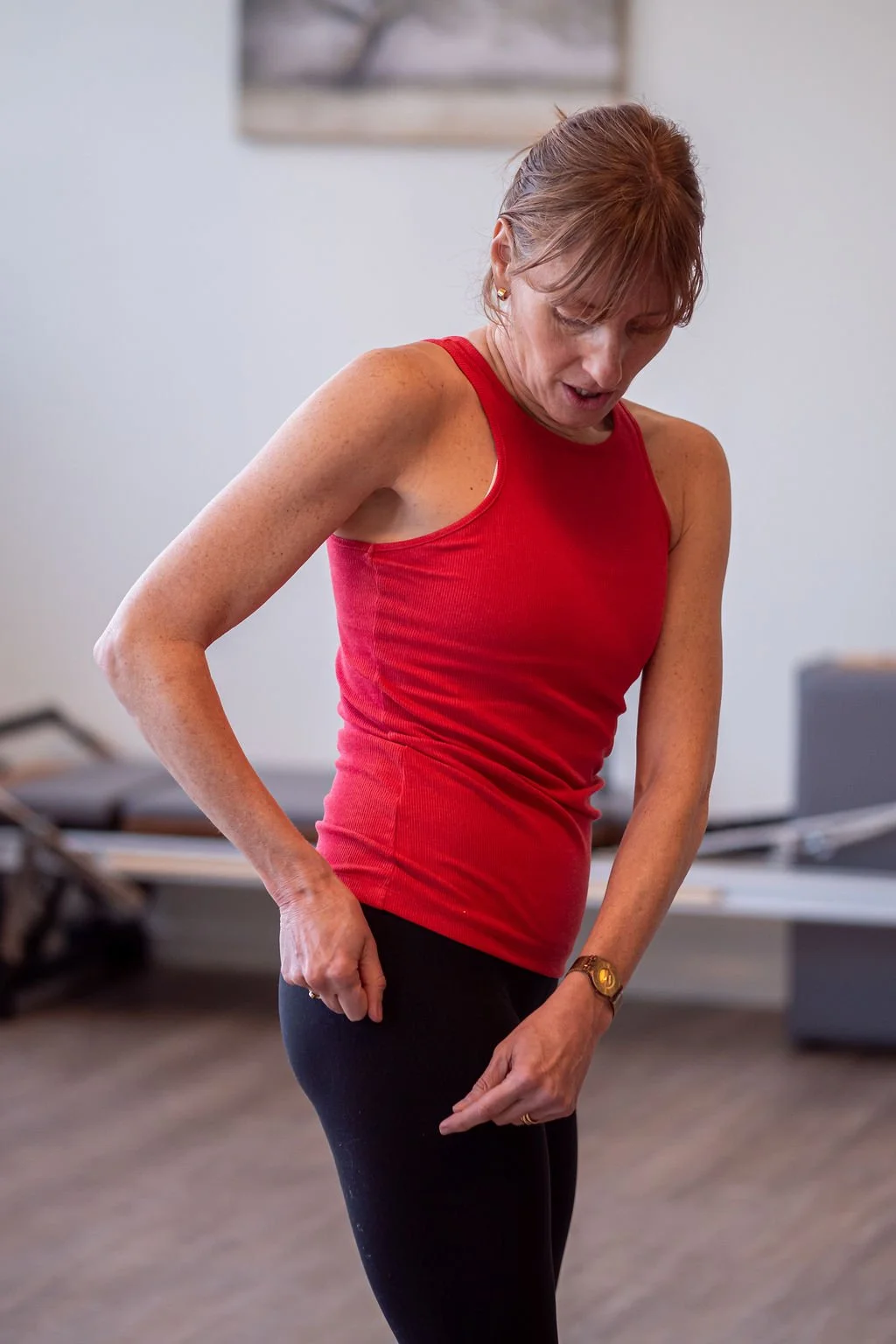 A woman with short reddish-brown hair in a red sleeveless top and black pants stands indoors, looking down and adjusting her pants.