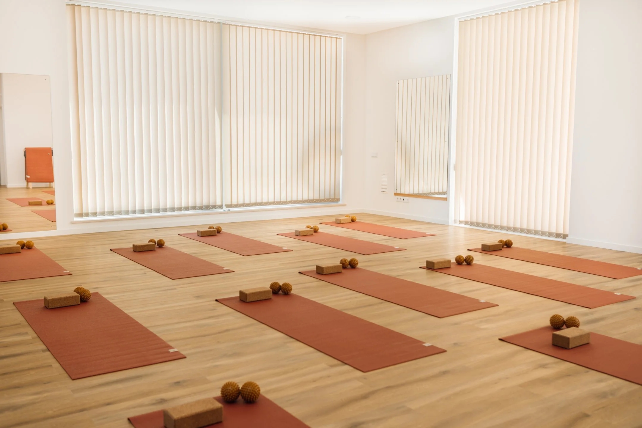 A yoga or meditation studio with wooden floors and large windows covered by vertical blinds. There are multiple orange yoga mats laid out in rows, each with a cork yoga block and a textured rubber ball nearby.