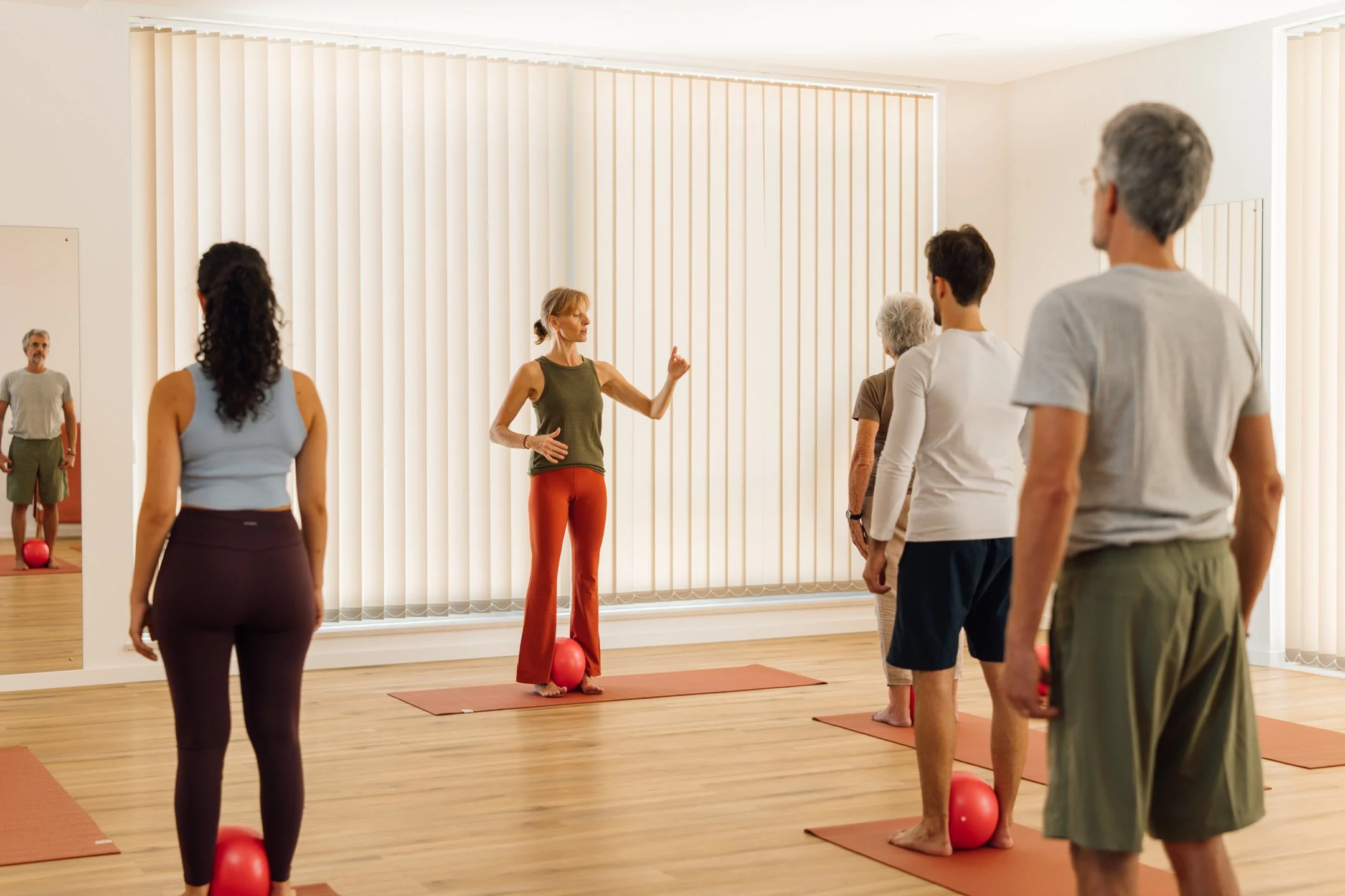 A yoga instructor leading a class of diverse people in a yoga studio with bamboo blinds, mats, and red exercise balls.