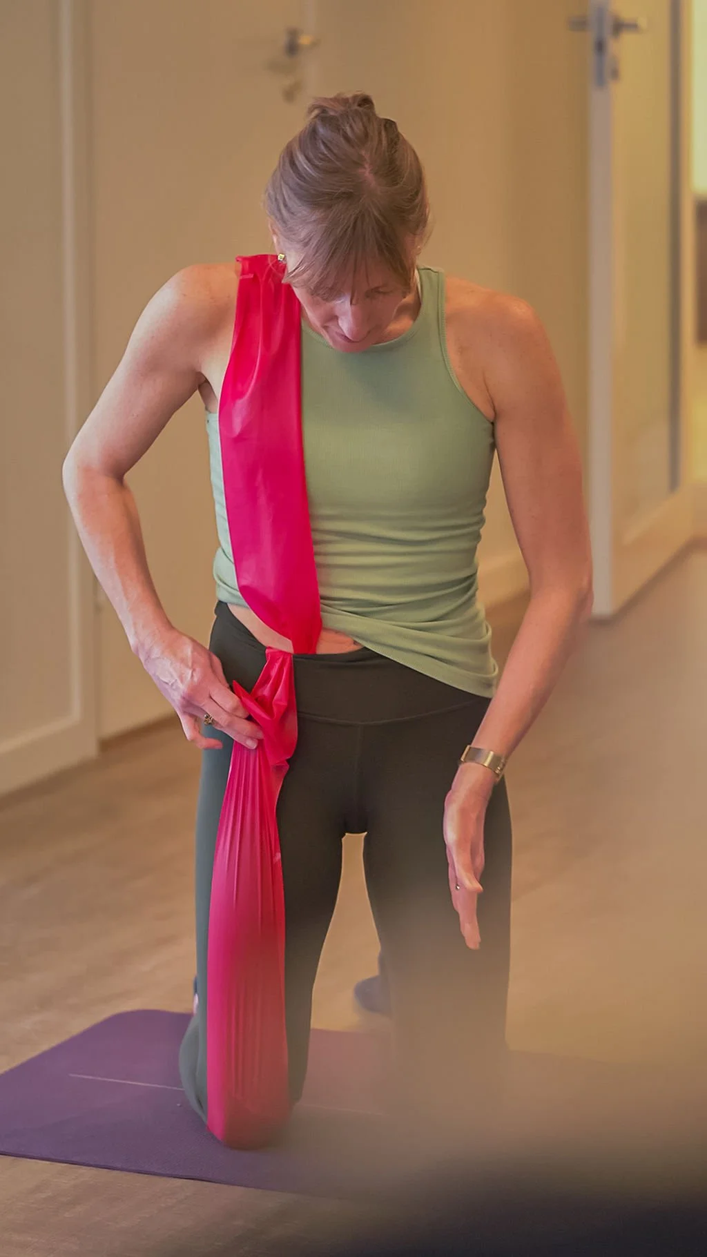 A woman in a green tank top and black leggings practicing yoga with a red yoga strap hanging from her shoulder, standing on a purple yoga mat in a room with wooden flooring.