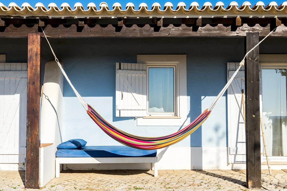 A colorful hammock with a blue pillow hanging between two dark wooden posts outside a blue house with white shutters and a window.