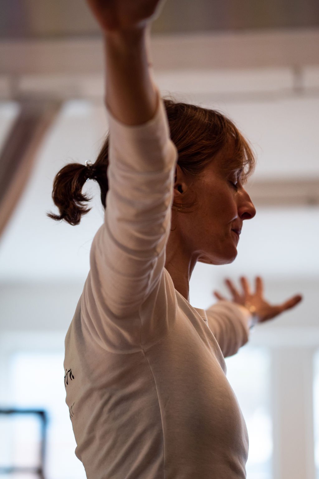 A woman with brown hair tied in a ponytail practicing yoga indoors, facing sideways with arms extended outward, eyes closed, and a peaceful expression.