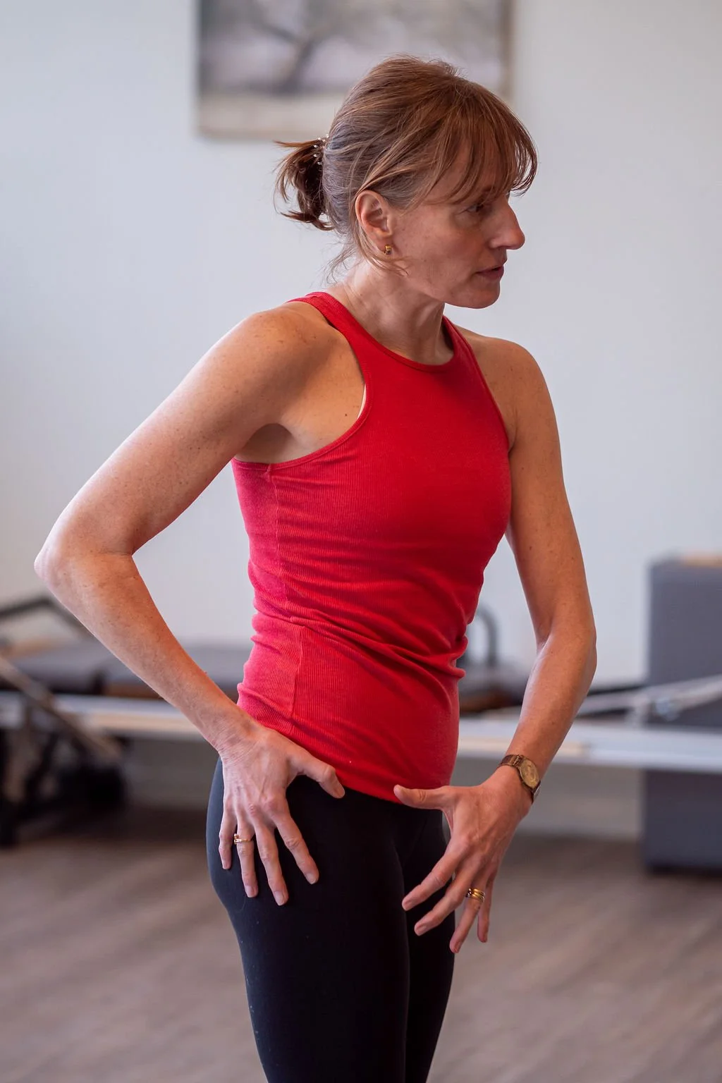 A woman in a red sleeveless top and black leggings stands with her hands on her hips, looking to the side, in a fitness or yoga studio.