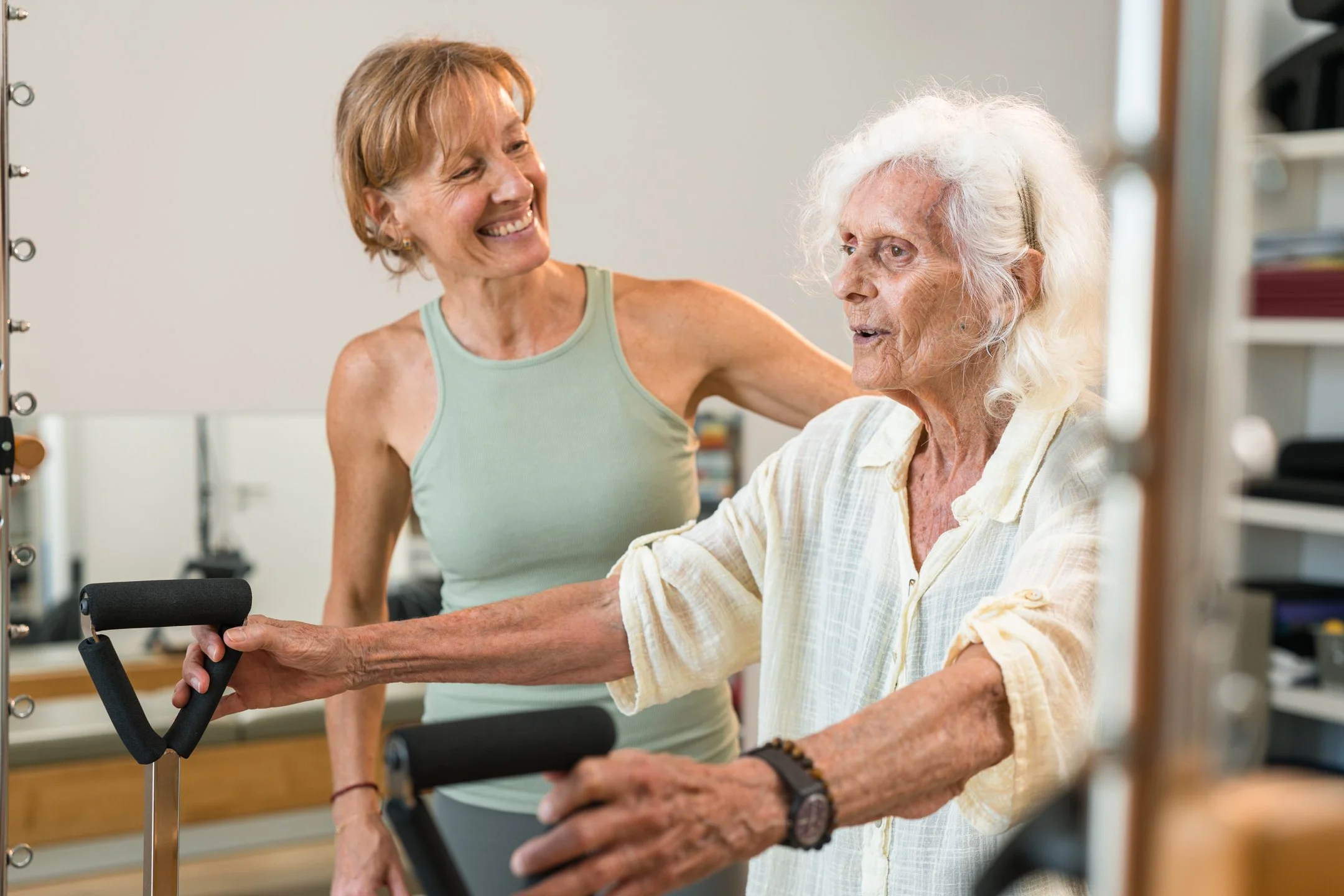 A young woman with short red hair smiling and helping an elderly woman with gray hair in a care or rehabilitation setting. The elderly woman is using a walker and appears to be engaging in a physical activity.