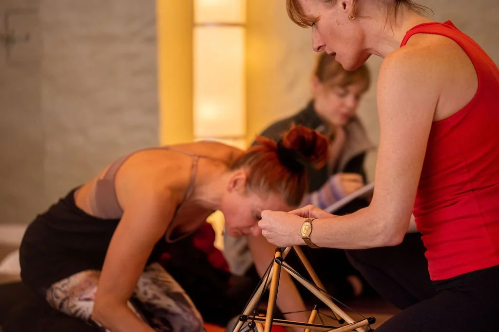 Two women engaged in a discussion, with one woman leaning forward on her knees and the other standing, holding a tripod or stand, in a warmly lit indoor setting. A third person is visible in the background, sitting and looking down.