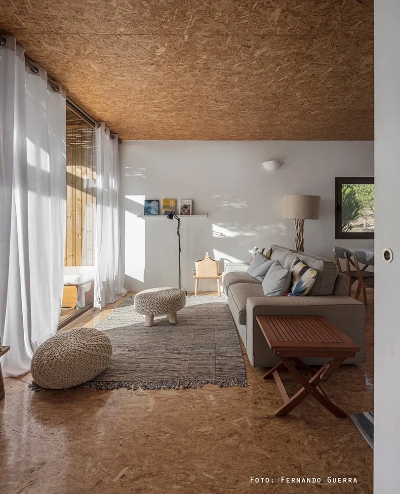 Bright living room with white walls, wooden ceiling, white curtains, and a large window. Contains a beige sofa with multiple pillows, a floor lamp, a small wooden table, a textured ottoman, a small chair, a rug, and framed pictures on a shelf. Sunlig