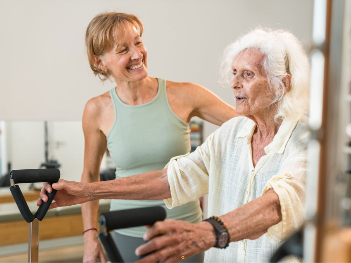 A woman happily assisting an elderly woman with walking exercises using a walker in a gym or rehabilitation center.