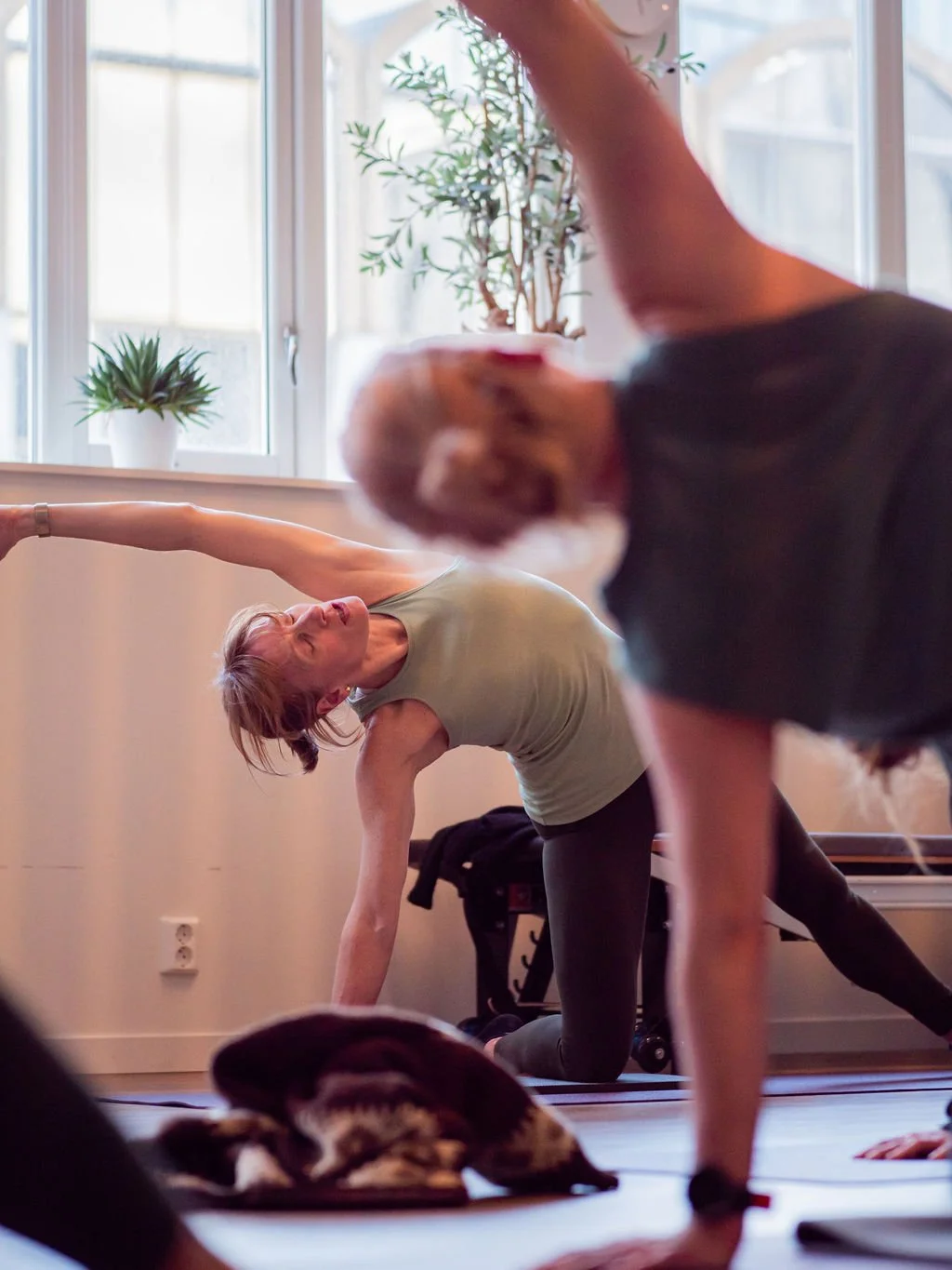 People practicing yoga in a room with large windows and potted plants, focusing on stretching and balance.