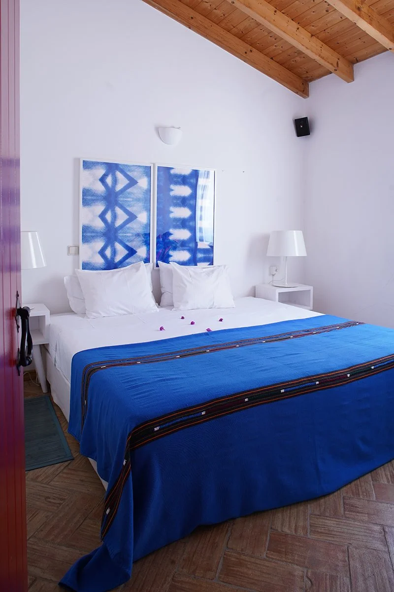 A neatly made bed with white pillows and a blue bedspread, in a room with white walls, wooden ceiling beams, and two white bedside tables with lamps. Two blue and white artwork pieces are hanging above the bed.