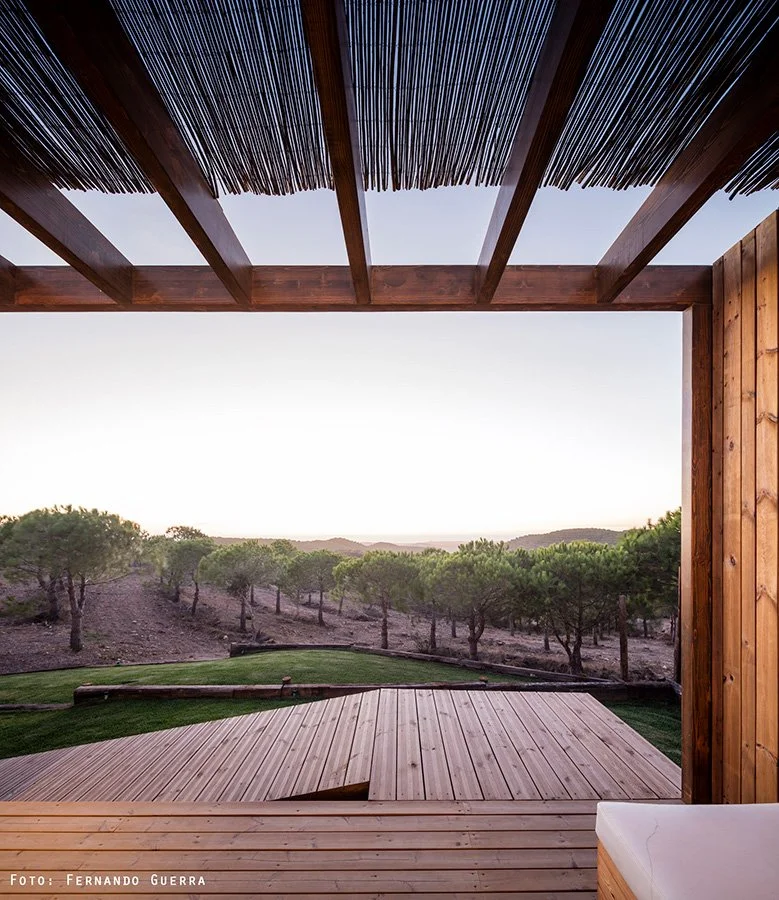View from a wooden deck overlooking a landscape with trees and hills at sunset, with a wooden ceiling and partial roof structure in the foreground.