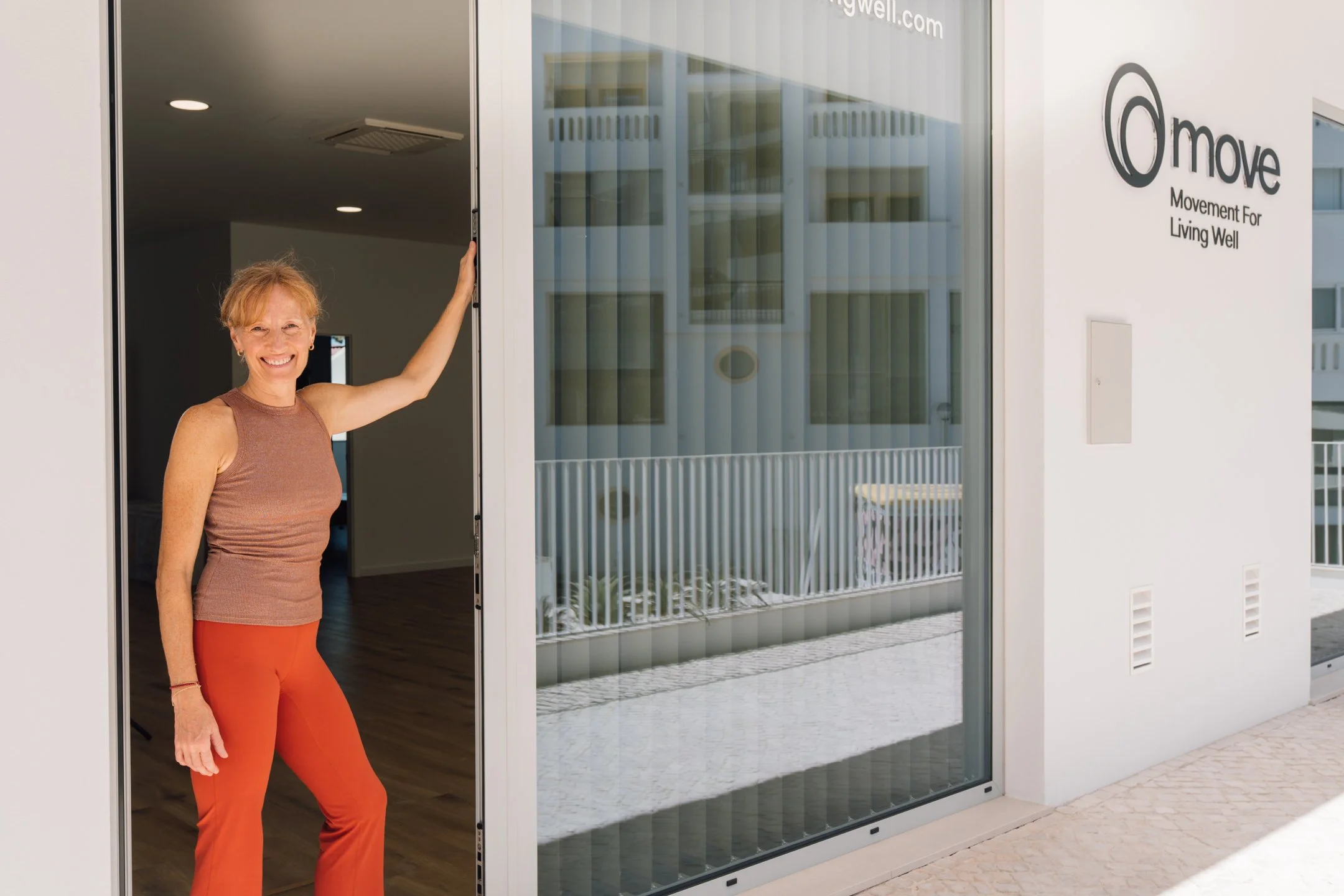 A woman stands smiling in a doorway, holding the door frame, inside a modern building with wooden flooring and a minimalist interior.