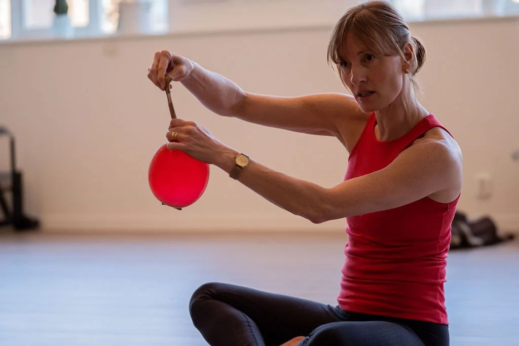 A woman in a red tank top and black leggings inflates a red balloon indoors.