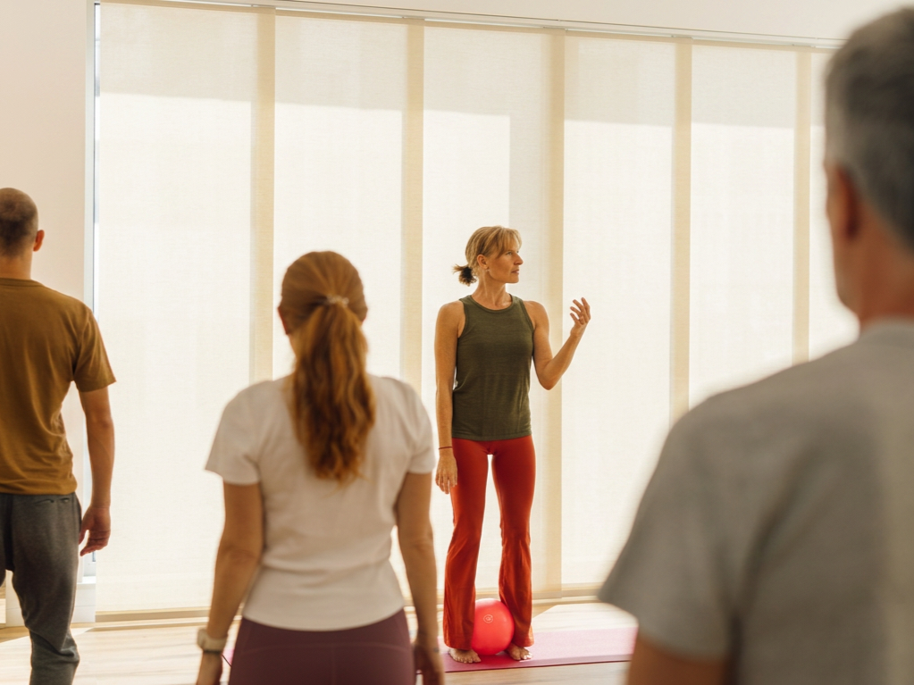 A yoga instructor demonstrates a pose to a group participating in a yoga class in a bright studio with large windows, using a pink yoga mat and a pink exercise ball nearby.