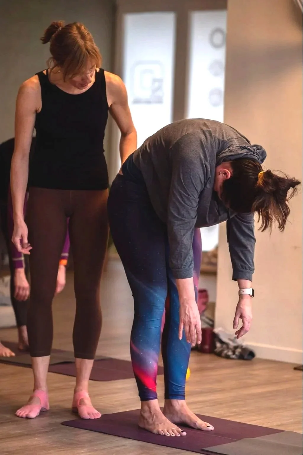 Two women practicing yoga indoors, one in a black tank top and leggings, the other in a gray hoodie and galaxy-patterned leggings, performing a forward bend on yoga mats.
