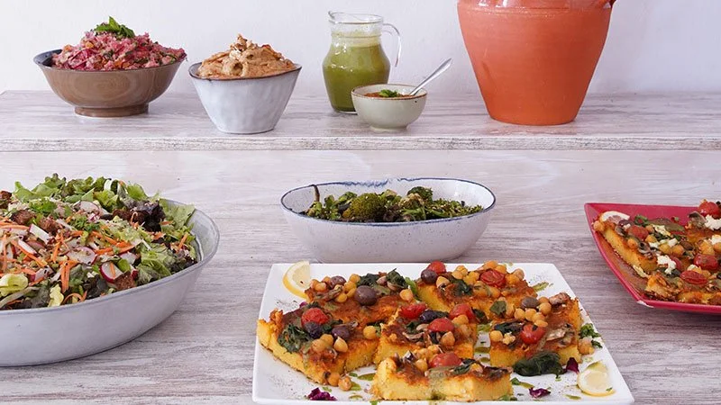 Assorted dishes including a vegetable salad, a dish with broccoli, a vegetable pizza with toppings like corn and tomatoes, and a pie or quiche on a light-colored wooden table.