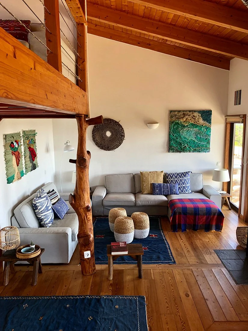 Living room with wooden ceiling and flooring, two sofas with decorative pillows, a small wooden coffee table, colorful woven ottomans, and wall art including a circular woven piece and a landscape painting.