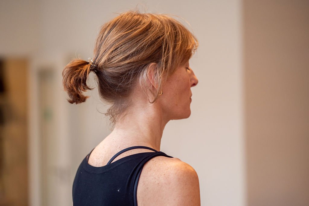 Side profile of a woman with red hair tied back in a ponytail, wearing a black tank top, standing indoors.