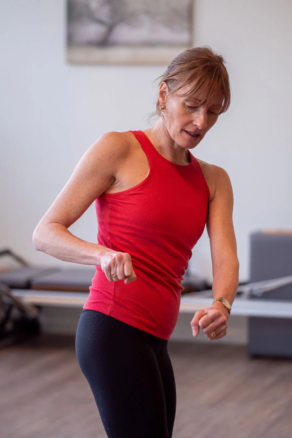 A middle-aged woman with reddish-brown hair in a ponytail, wearing a red sleeveless top and black leggings, exercising indoors.