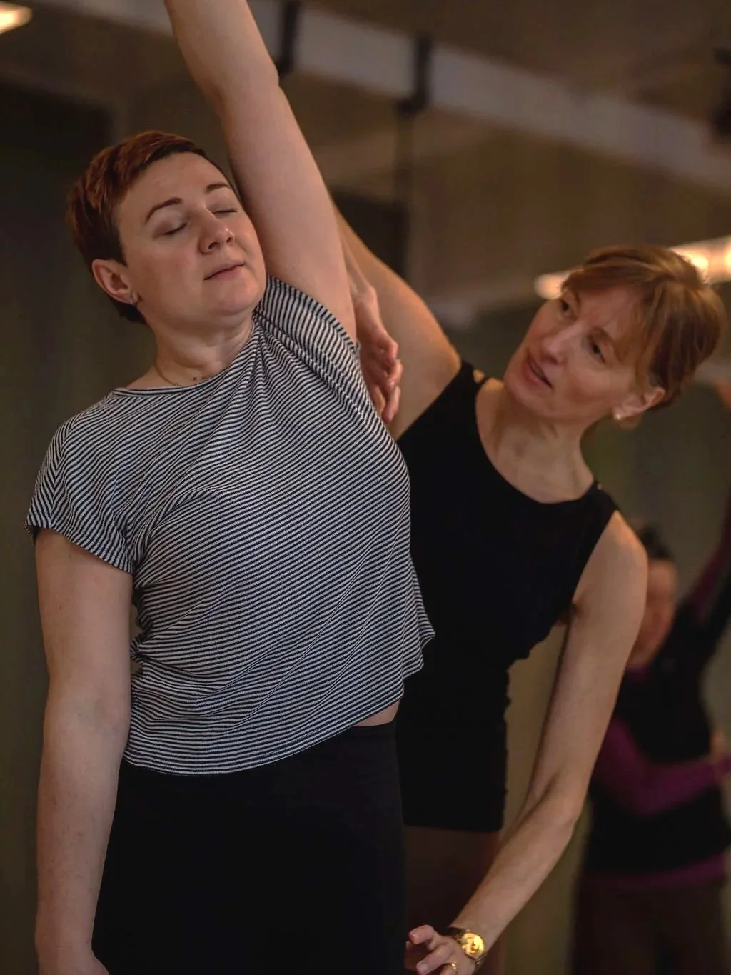 Two women practicing yoga together, one with short reddish hair wearing a striped shirt and the other with short light brown hair wearing a black top, in an indoor yoga studio.