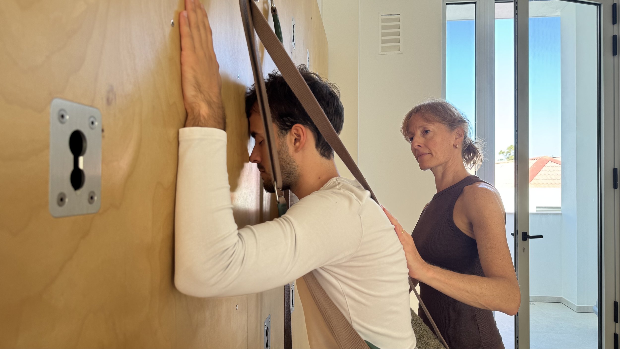 A young man is participating in a harness-based activity on a wooden wall, leaning forward with his forehead touching the surface, while a woman appears to be a trainer or assistant providing support or instructions.