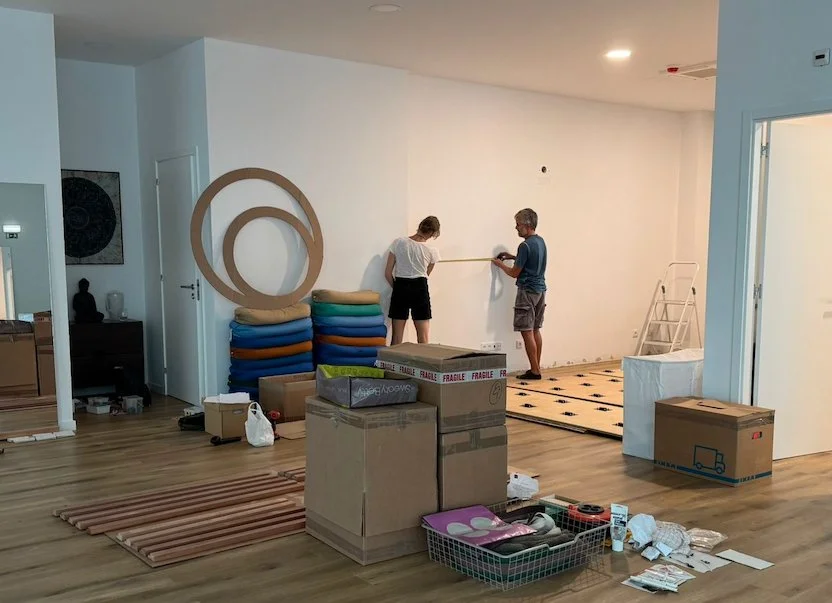 Two people are measuring and preparing to paint a white wall in a living room filled with moving boxes and packed supplies, with fitness mats and yoga equipment stored nearby.