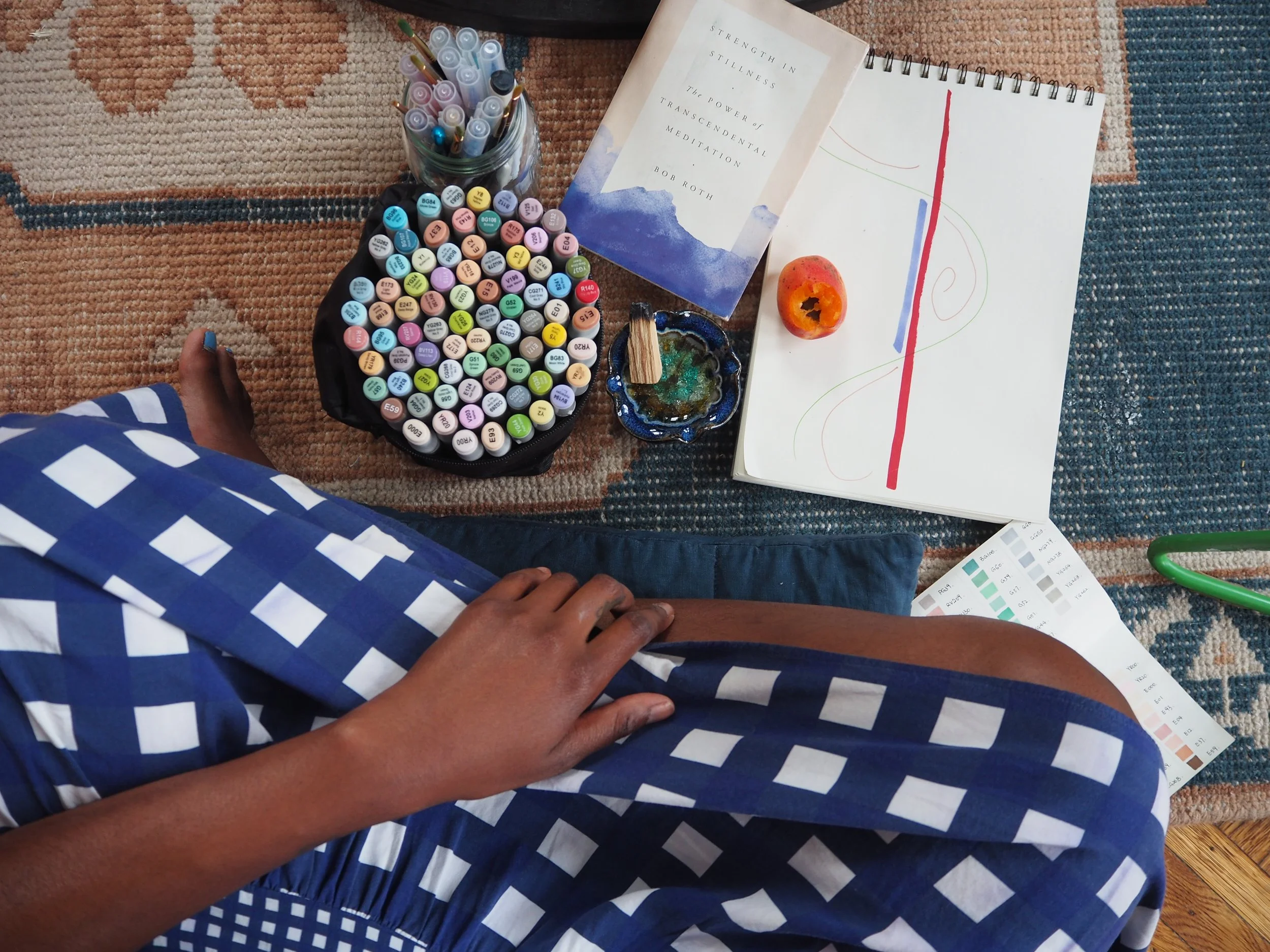 A creative workspace on a patterned rug with colored markers, notebooks, color swatches, fruit, and the book Strength in Stillness.