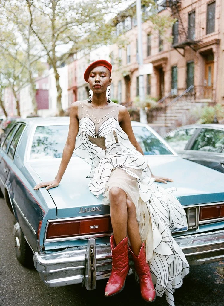 A woman in a floral dress, red boots, and a red beret sits on the back of a blue vintage car on a tree-lined Brooklyn street.