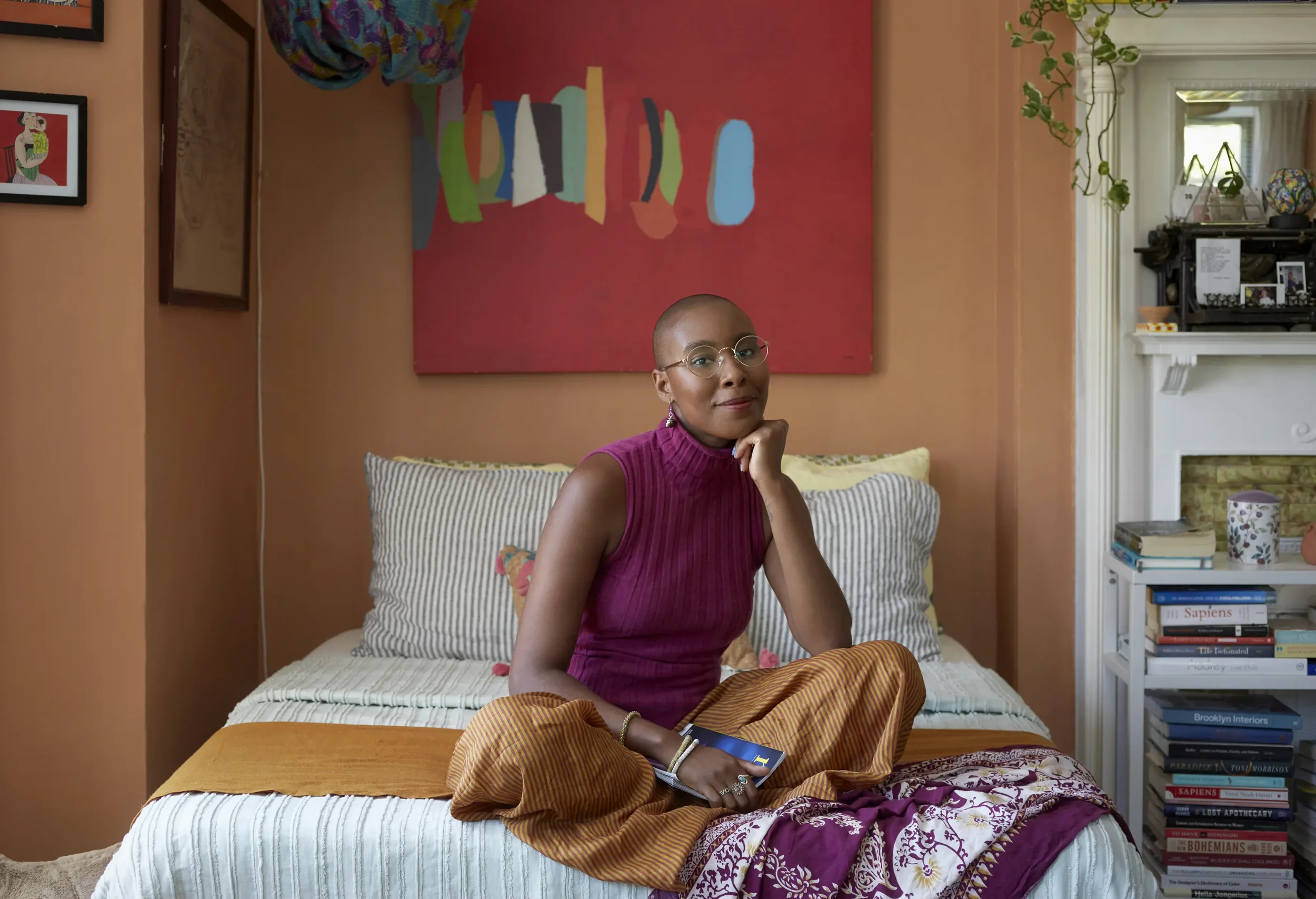A woman with glasses and a shaved head sits on a bed in a colorful bedroom with posters and bookshelves behind her.