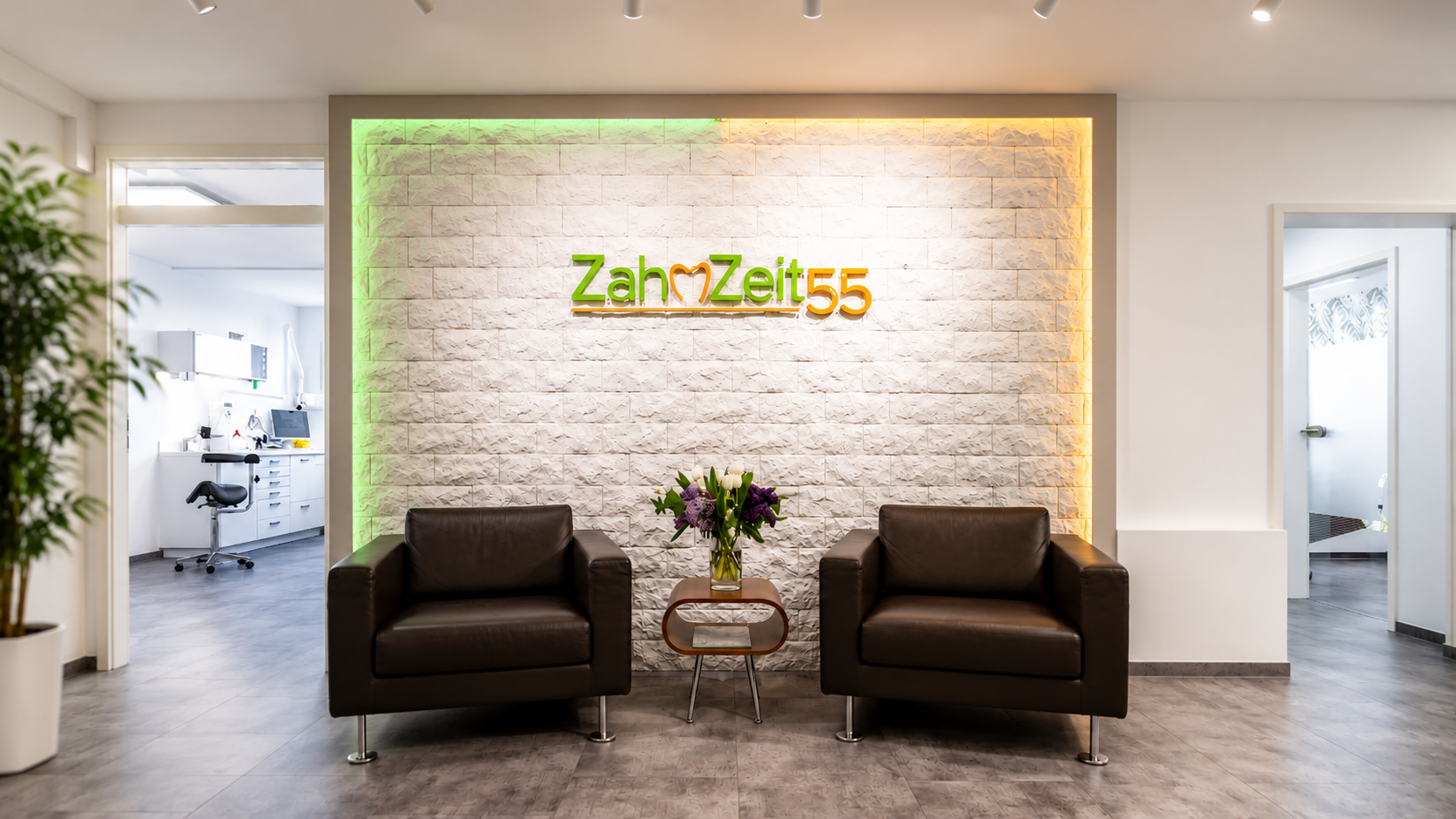 Wartebereich in einer Zahnarztpraxis mit zwei schwarzen Ledersofas, einem Beistelltisch mit Blumen, einer weißen Wand mit buntem Praxislogo "ZahnZeit55" und zwei offene Türen zu Behandlungsräumen.