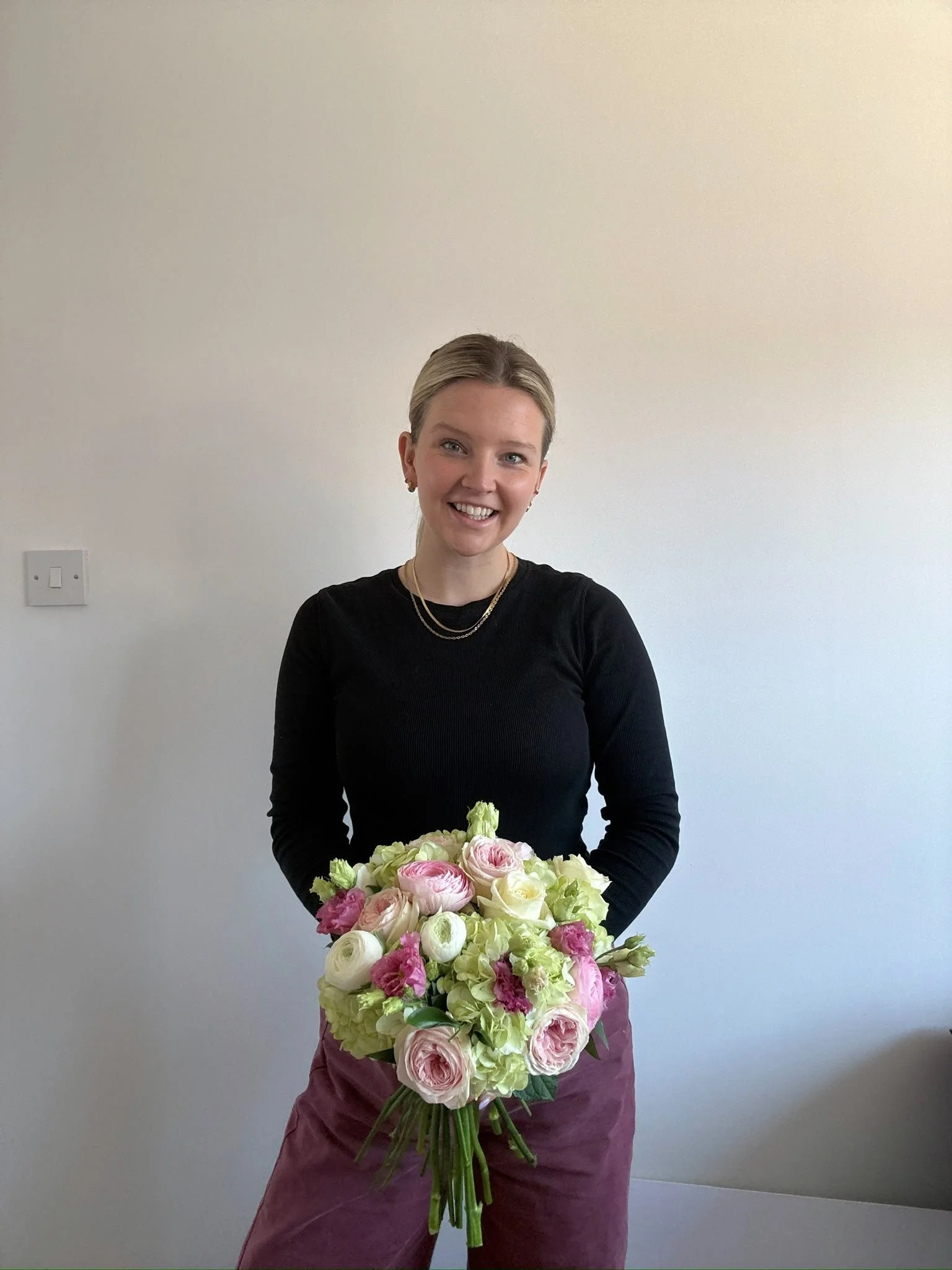 A woman with blonde hair wearing a black top and gold jewelry holding a large bouquet of pink and white flowers in front of a plain light-colored wall.