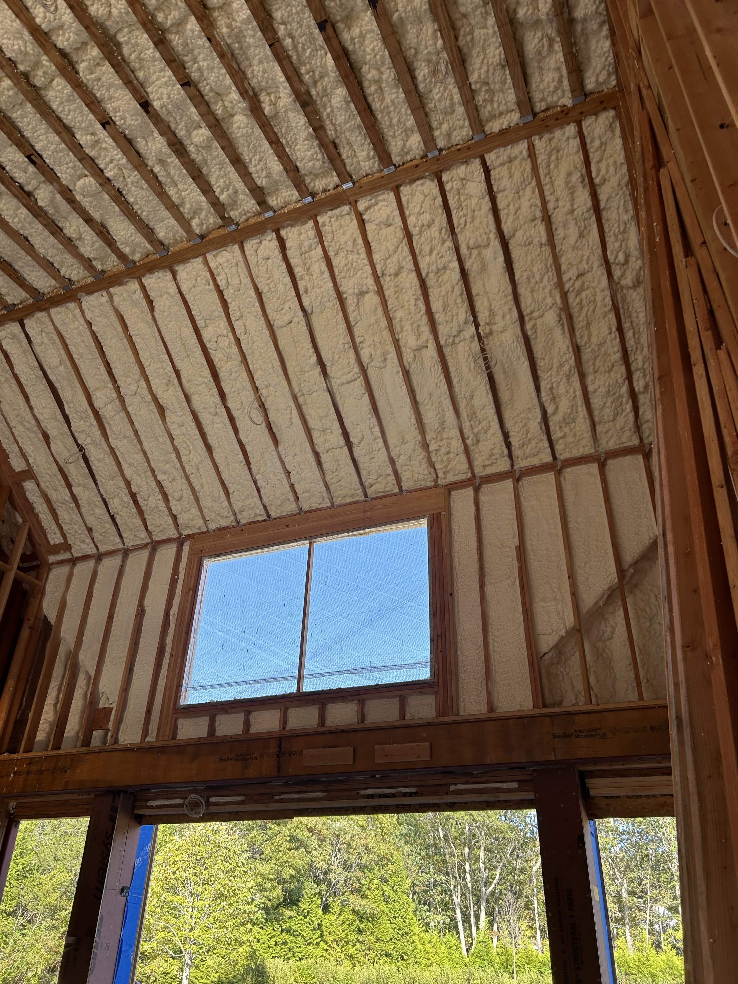 Interior view of a house under construction, showing roof trusses and insulation, with a large window looking out to a wooded area.