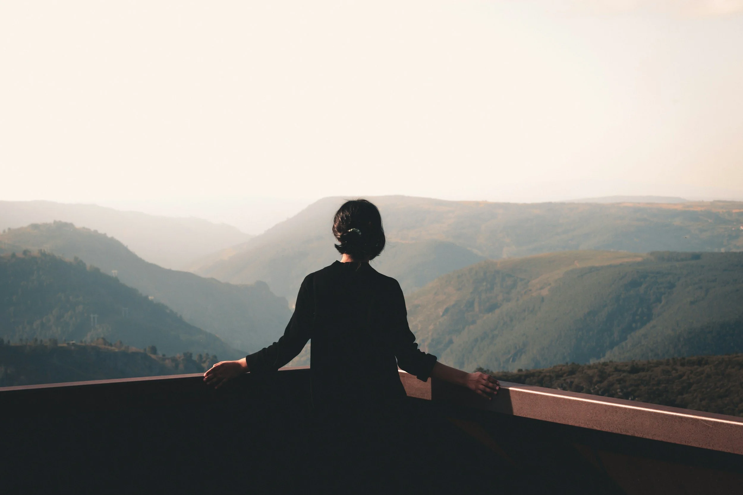 A woman practicing meditation or yoga on a balcony overlooking a scenic mountainous landscape during sunset.