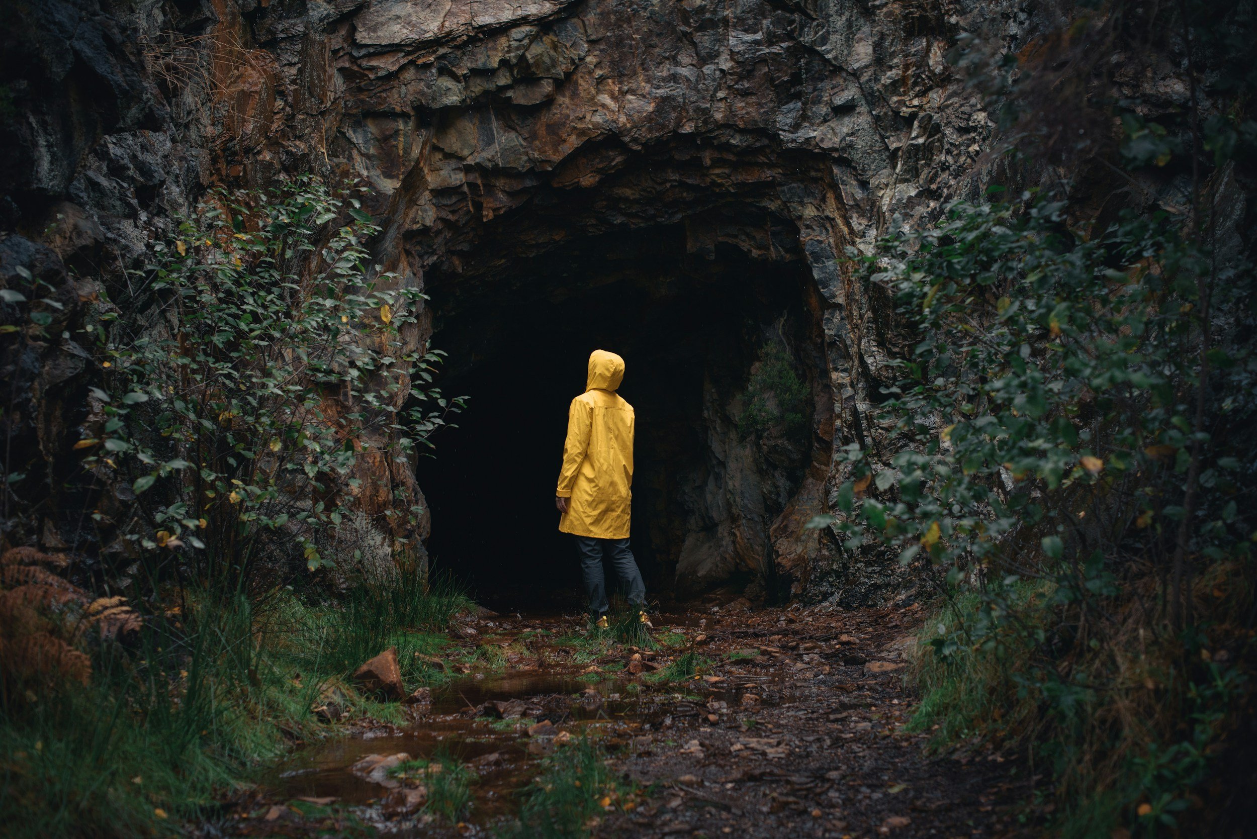 Person in a yellow raincoat standing in front of a dark cave entrance surrounded by rocks and greenery during rainy weather.