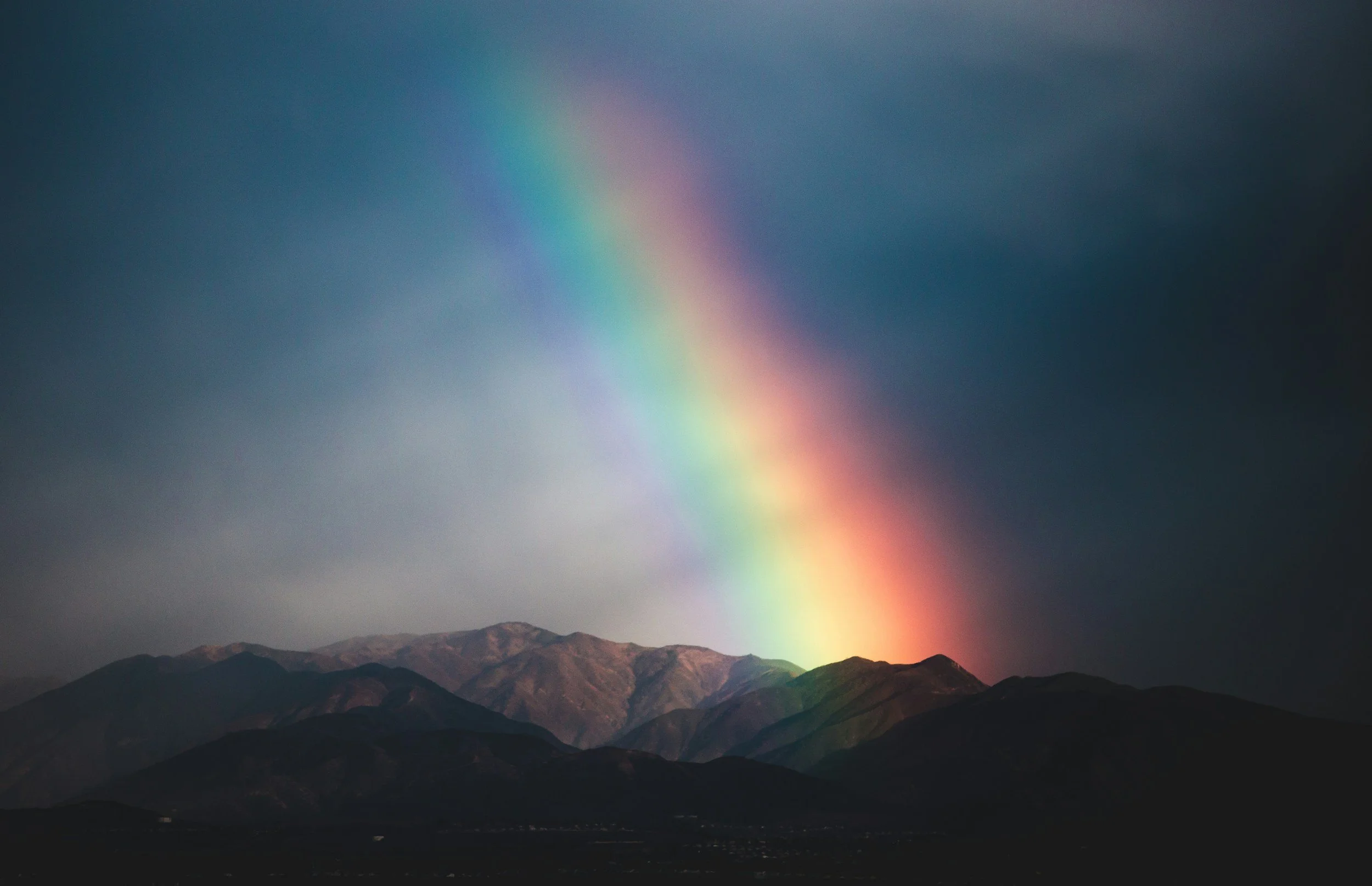 A rainbow arches over a mountain range with dark clouds in the sky.