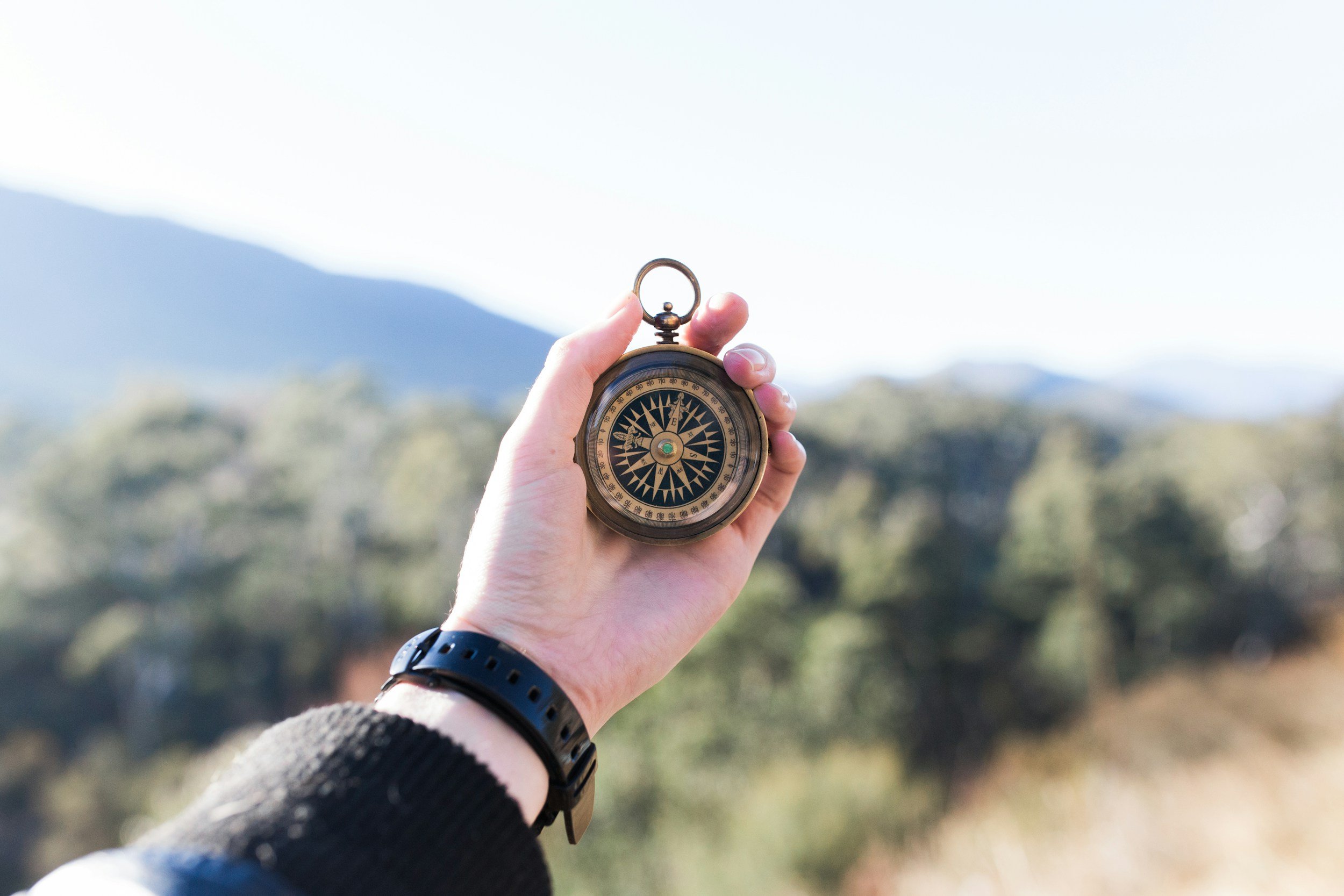 A hand holding a vintage compass outdoors with blurred trees and mountains in the background.
